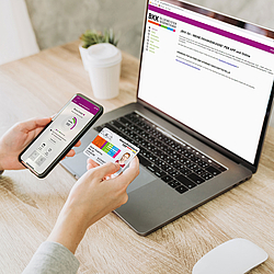 Eine Person sitzt vor einem Laptop. Auf dem Bildschirm ist die Internetseite der BKK GS. In Ihren Händen hält sie ein Smartphone und die Versichertenkarte.