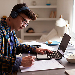 Ein Junge im Teenageralter sitzt an einem Schreibtisch. Er trägt große Kopfhörer. Links vor ihm steht ein Laptop, rechts liegt ein Block auf dem er etwas notiert.
