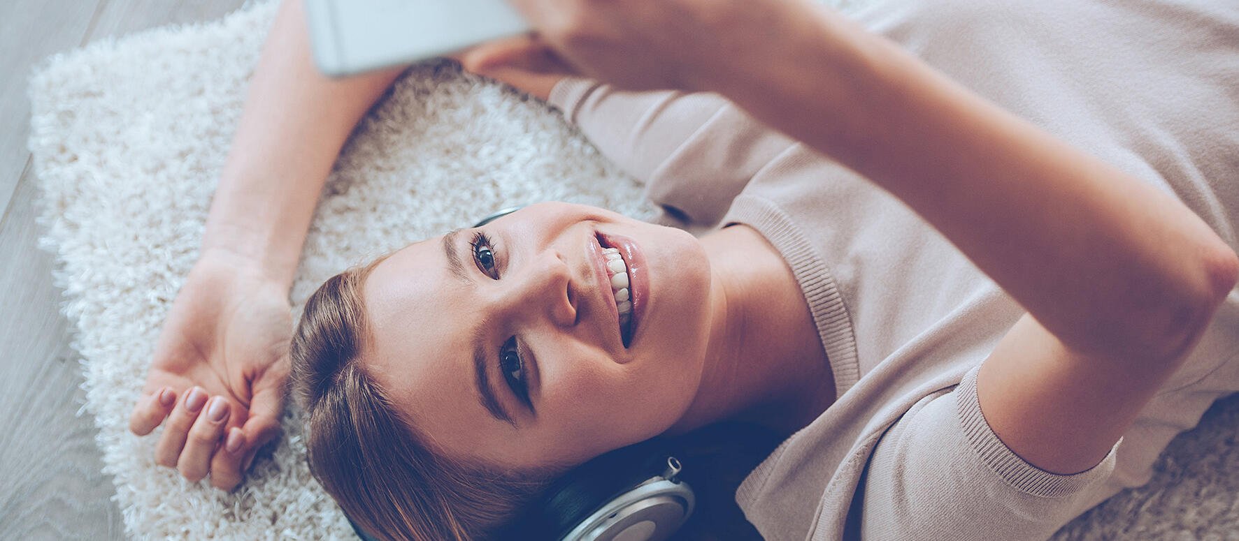 Junge Frau liegt auf Teppich mit Kopfhörern und schaut glücklich auf Smartphone