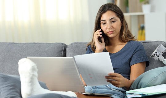 Frau telefoniert mit Gipsfuß auf dem Sofa am Laptop 