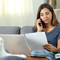 Eine junge Frau sitzt auf dem Sofa und hat ihr gebrochenes Bein hochgelegt. Sie hat einen Laptop auf dem Schoss, Unterlagen in der Hand und telefoniert.