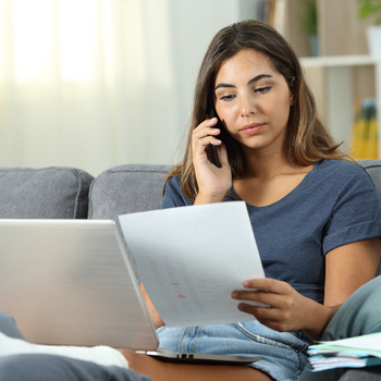 Eine junge Frau sitzt auf dem Sofa und hat ihr gebrochenes Bein hochgelegt. Sie hat einen Laptop auf dem Schoss, Unterlagen in der Hand und telefoniert mit einem Arzt, um sich eine Zweitmeinung einzuholen.