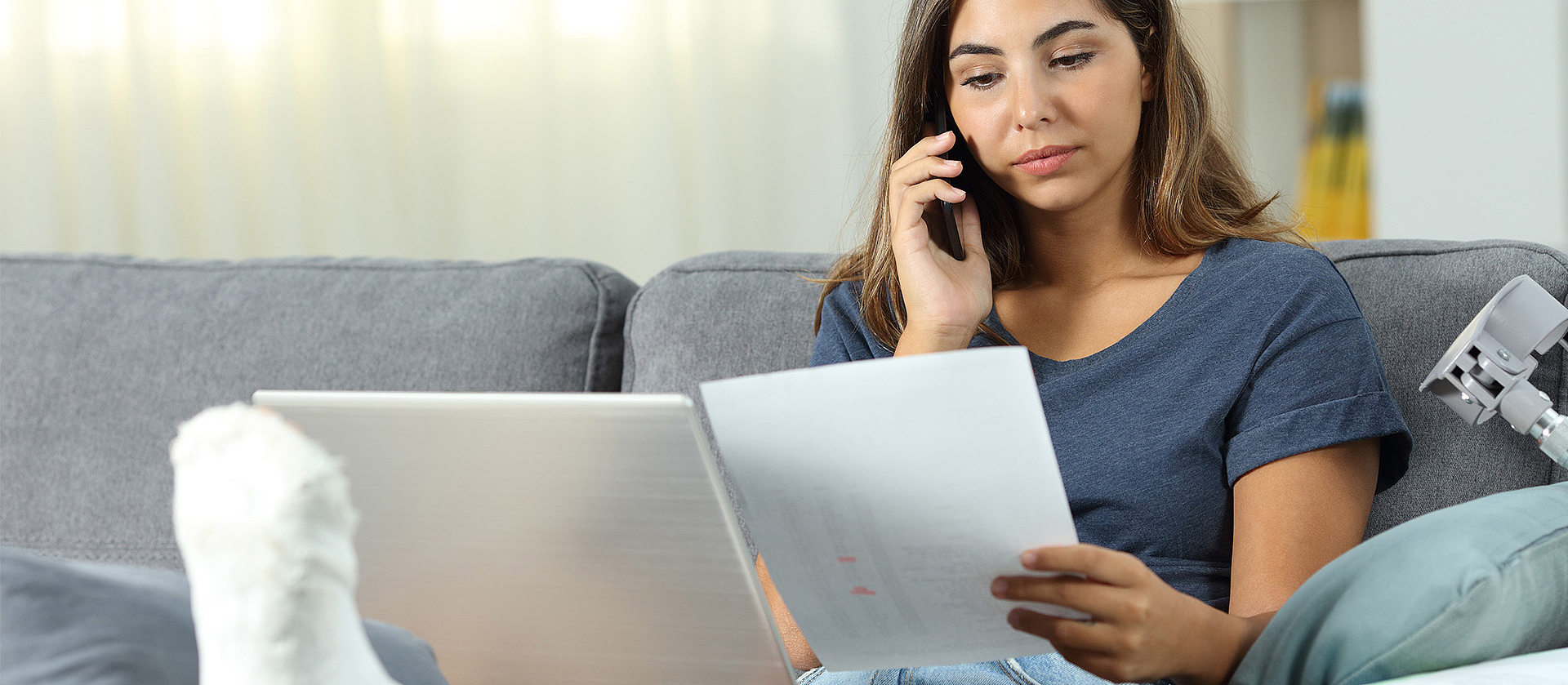 Eine junge Frau sitzt auf dem Sofa und hat ihr gebrochenes Bein hochgelegt. Sie hat einen Laptop auf dem Schoss, Unterlagen in der Hand und telefoniert.