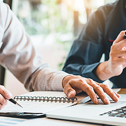 Zwei Männer sitzen an einem Tisch, vor ihnen steht ein Laptop. Einer schaut auf sein Smartphone, der andere schreibt in ein Notizbuch.