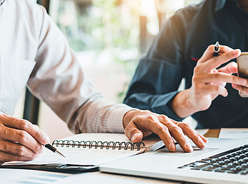 Zwei Männer sitzen an einem Tisch, vor ihnen steht ein Laptop. Einer schaut auf sein Smartphone, der andere schreibt in ein Notizbuch.