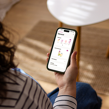 Eine Frau mit lockigen Haaren sitzt in ihrem Wohnzimmer. Auf ihrem Smartphone nutzt sie die ePA App.