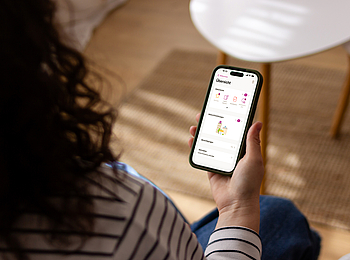 Eine Frau mit lockigen Haaren sitzt in ihrem Wohnzimmer. Auf ihrem Smartphone nutzt sie die ePA App.
