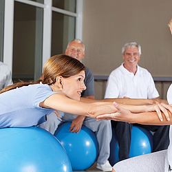 Eine junge Frau mit langem Haar liegt während eine Physiotherapiestunde auf einem Gymnastikball. Die Therapeutin hält sie an den Armen festhält. 