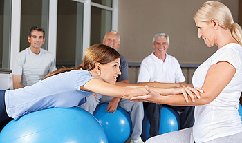 Eine junge Frau mit langem Haar liegt während eine Physiotherapiestunde auf einem Gymnastikball. Die Therapeutin hält sie an den Armen festhält. 