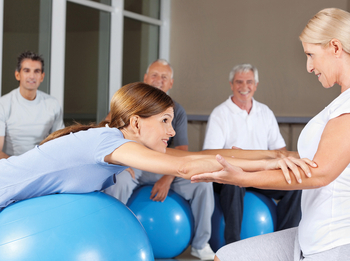 Eine junge Frau mit langem Haar liegt während eine Physiotherapiestunde mit der Brust auf einem Gymnastikball. Sie schaut ihre Therapeutin an, die sie an den Armen festhält. Im Hintergrund sitzen weitere Patienten und schauen Ihnen zu.