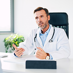 Ein Arzt sitzt an seinem Schreibtisch und unterhält sich mit seinen Patienten.