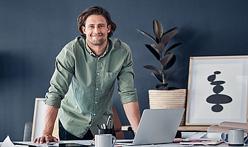 Ein junger Mann mit längeren braunen Haaren und kurzem Bart schaut lachend in die Kamera. Er steht in einem Büro und stützt sich mit den Händen auf einem Schreibtisch ab. Auf dem Tisch stehen ein Laptop eine Kaffetasse und andere Büroutensilien. 