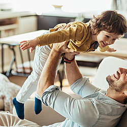 Ein junger Vater mit Bart und dunklen Haaren sitzt auf einem Sessel. Er hebt seinen Sohn hoch. Der kleine Junge lacht und breitet die Arme.