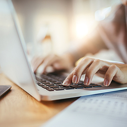 Eine Frau sitzt an einem Schreibtisch. Sie schreibt etwas an einem Laptop. 