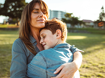 Eine Frau mit langen Haaren umarmt ihren Sohn. Sie stehen bei Sonnenschein auf einer Wiese.