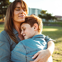 Eine Frau mit langen Haaren umarmt ihren Sohn. Sie stehen bei Sonnenschein auf einer Wiese.