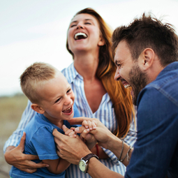 Ein junger Vater mit Bart lacht ausgelassen und kitztelt seinen kleine blonden Sohn am Hals. Die Mutter lacht ebenfalls und hält den kleinen Jungen im Arm. 