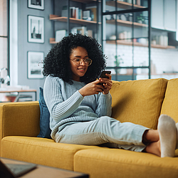 Eine Frau mit lockigen Haaren und Brille sitzt auf einem gelben Sofa in. Sie schaut lächelnd auf ein Smartphone.