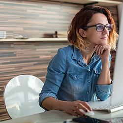Eine Frau mit roten Haaren und Brille sitzt an einem Schreibtisch. Sie schaut auf den Monitor eines Computers. 