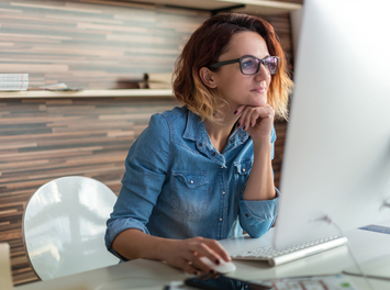 Eine junge Frau mit schulterlangen roten Haaren und Brille sitzt an einem Schreibtisch. Sie schaut auf den Monitor eines Computers. Mit der rechten Hand bedient sie den PC, auf die rechte hat sie ihr Kinn aufgestützt.