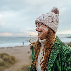 Eine rothaarige Frau steht am Strand und lächelt. Sie trägt eine Mütze und ein grüne Jacke.