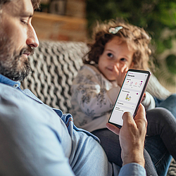 Mann und Tochter sitzen auf dem Sofa. Der Mann schaut in die ePA-App auf seinem Smartphone.