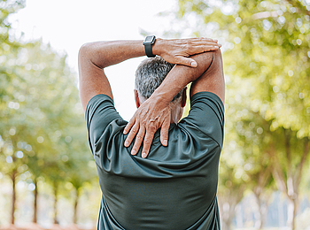 Ein Mann steht in der Natur und dehnt seinen Schulterbereich. Im Hintergrund stehen Bäume.