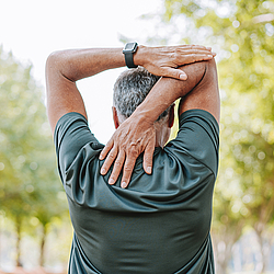 Ein Mann steht in der Natur und dehnt seinen Schulterbereich. Im Hintergrund stehen Bäume.