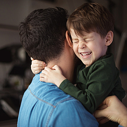 Ein Vater mit dunklen kurzen Haaren umarmt seinen kleinen Sohn. Der Junge kneift die Augen zusammen und lacht ausgelassen.