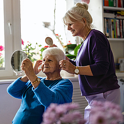Eine blonde Frau steht hinter ihrer Mutter und macht ihr die Haare. 