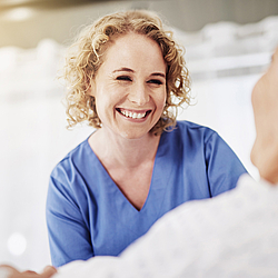 Eine Krankenschwester mit blonden lockigen Haaren trägt einen blauen Kittel. Sie lächelt eine ältere Patientin im Pflegebett an.
