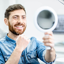 Ein Mann sitzt auf einem Behandlungsstuhl beim Zahnarzt. Er hält einen Spiegel in der Hand und betrachtet seine Zähne.
