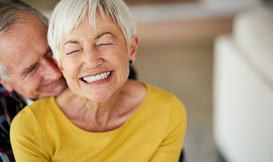 Eine ältere Frau mir kurzen weißen Haaren lacht in die Kamera. Ein älterer Mann umarmt sie und legt den Kopf auf ihre Schulter.