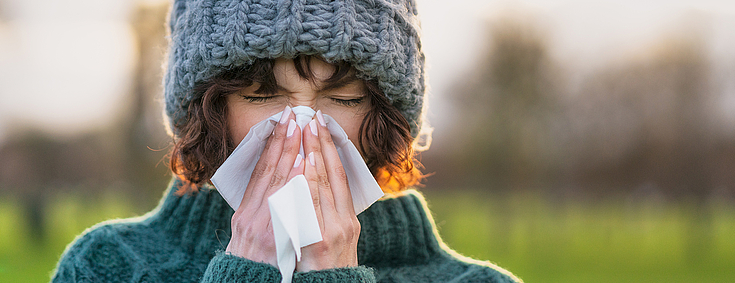 Eine junge Frau mit kurzen lockigen Haaren schnäuzt in ein weißes Taschentuch. Sie hat die Augen geschlossen. Sie trägt eine graue Wolllmütze und einen grünen Strickpullover.