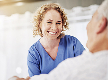 Eine Krankenschwester mit lockigen blonden Haaren schaut auf eine Patientin im Krankenhausbett und lacht sie fröhlich an.