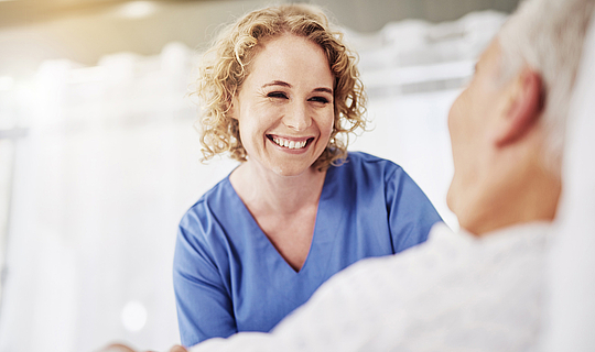 Eine Krankenschwester mit lockigen blonden Haaren schaut auf eine Patientin im Krankenhausbett und lacht sie fröhlich an.