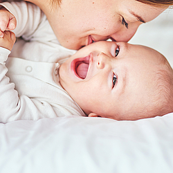 Ein Baby in einem weißen Body liegt auf einer Matratze und lächelt. Seine Mutter gibt ihm einen Kuss auf die Wange.