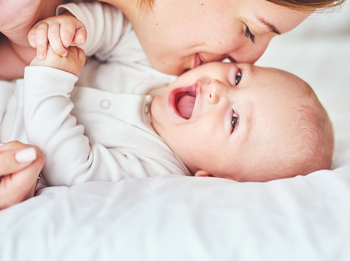 Ein Baby in einem weißen Body liegt auf einer Matratze. Es lacht fröhlich in die Kamera. Seine Mutter lächelt und gibt ihm einen Kuss auf die Wange.