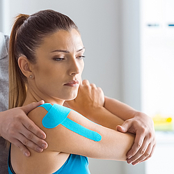 Ein Physiotherapeut führt bei einer Frau eine Schulterbehandlung durch. Die Patientin hat Kinesio-Tape auf Arm und Schulter.
