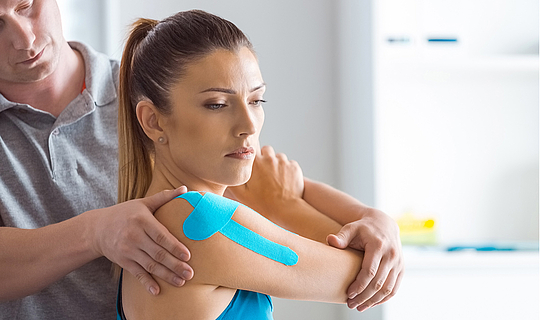 Ein Physiotherapeut führt bei einer Frau eine Schulterbehandlung durch. Die Patientin hat Kinesio-Tape auf Arm und Schulter.