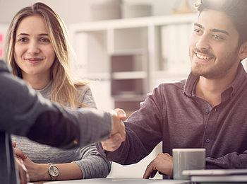Ein junges Paar sitzt in einem Beratungsgespräch, beide lächeln. Der Mann und der Berater geben sich die Hand.