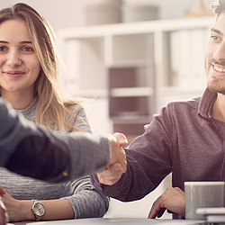 Ein junges Paar sitzt in einem Beratungsgespräch, beide lächeln. Der Mann und der Berater geben sich die Hand.