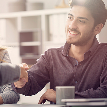 Ein junges Paar sitzt in einem Beratungsgespräch, beide lächeln. Der Mann und der Berater geben sich die Hand.