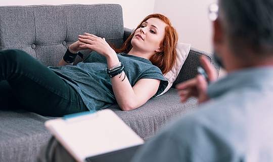Eine junge Frau mit langen roten Haaren liegt während ihrer psychotherapeutischen Behandlung auf einer grauen Couch.