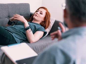 Eine junge Frau mit langen roten Haaren liegt während ihrer psychotherapeutischen Behandlung auf einer grauen Couch.