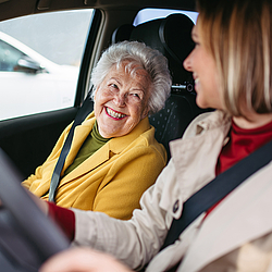 Eine ältere Frau mir kurzen grauen Haaren und eine junge Frau mit schulterlangen hellen Haaren sitzen in Auto und schauen sich an.