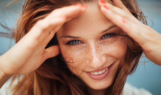 Eine Frau mit langen roten Haaren lächelt. Sie hält ihre Hände an ihr Gesicht, damit der Wind ihr keine Haare ins Gesicht weht.