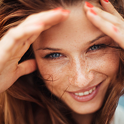Eine Frau mit langen roten Haaren lächelt. Sie hält ihre Hände an ihr Gesicht, damit der Wind ihr keine Haare ins Gesicht weht.