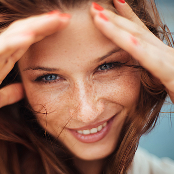 Eine Frau mit langen roten Haaren lächelt. Sie hält ihre Hände an ihr Gesicht, damit der Wind ihr keine Haare ins Gesicht weht.