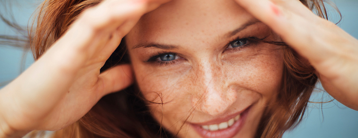 Eine junge FRau mit langen roten Haaren und Sommersprossen lächelt in die Kamera. Sie hält ihre Hände links und rechts an ihr Gesicht, damit der Wind ihr keine Haare ins Gesicht weht.